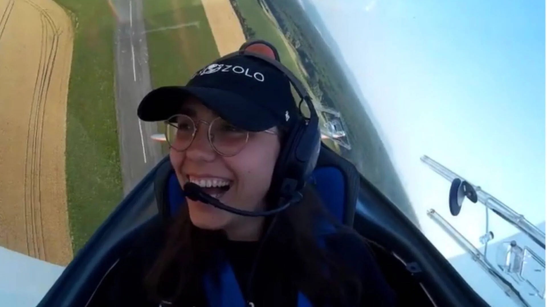 Joven con auriculares y micrófono sonríe dentro de la cabina de un planeador durante un vuelo, con campos verdes visibles abajo.