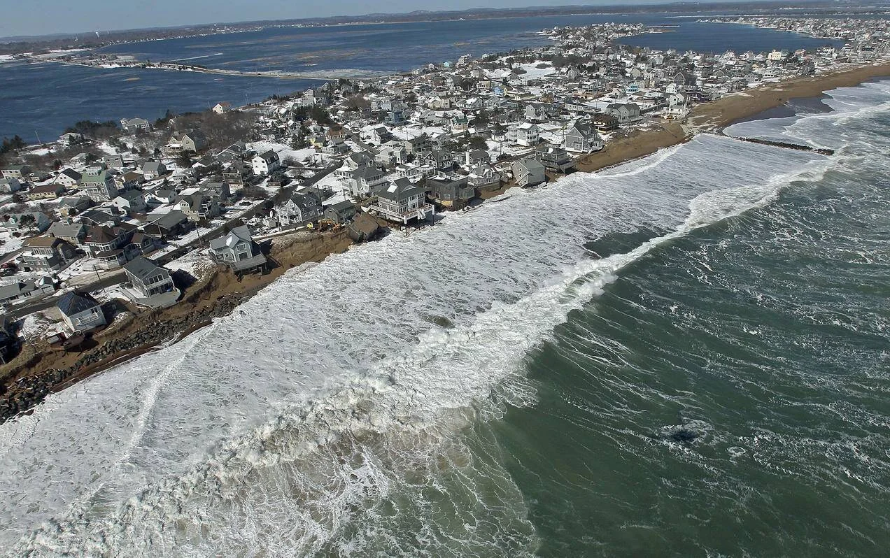Vista aérea de una localidad costera nevada con casas al borde de un acantilado erosionado por olas intensas del océano.