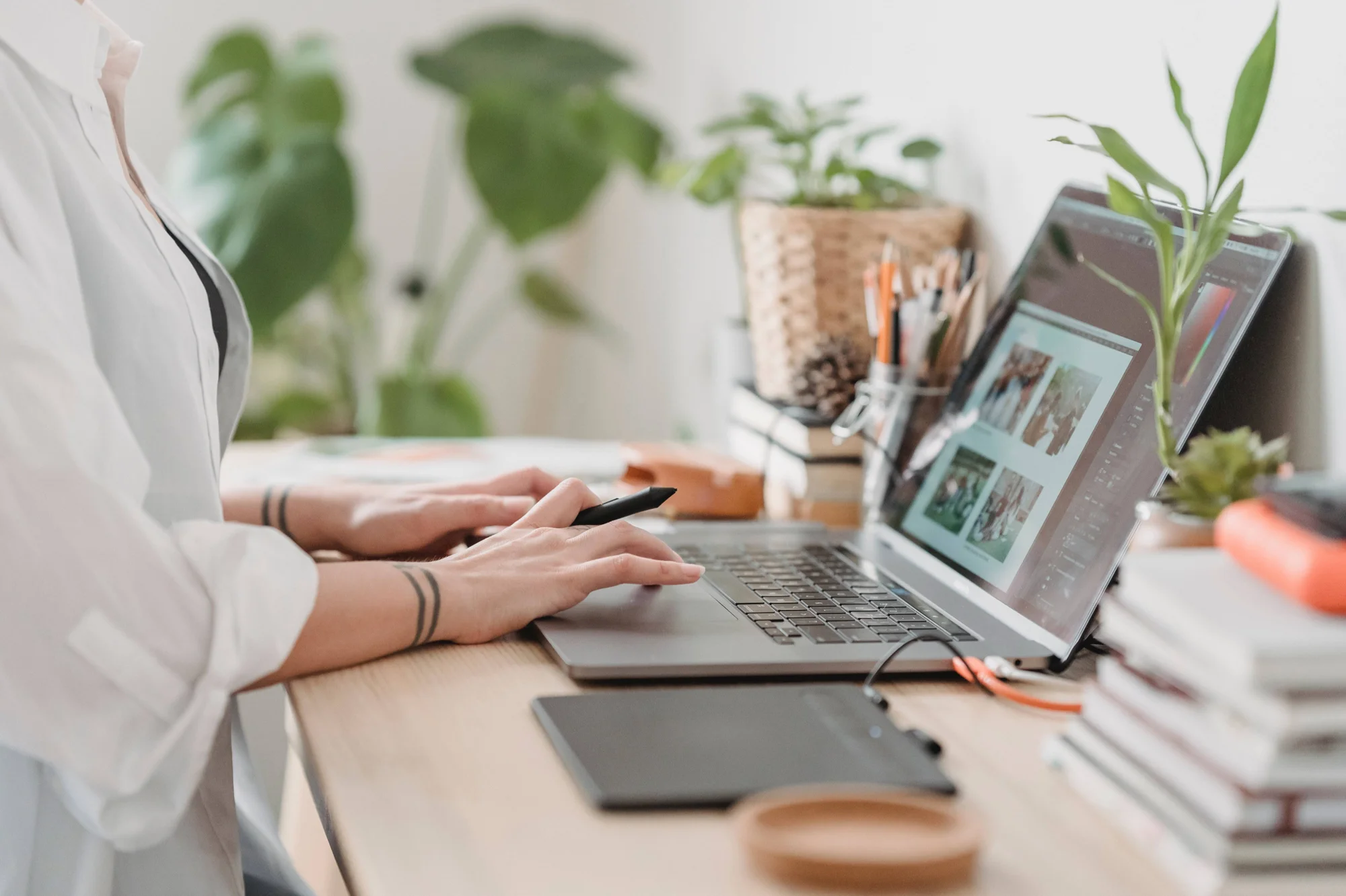 Persona edita imágenes en software de diseño usando una laptop y una tableta digitalizadora en un escritorio con plantas.