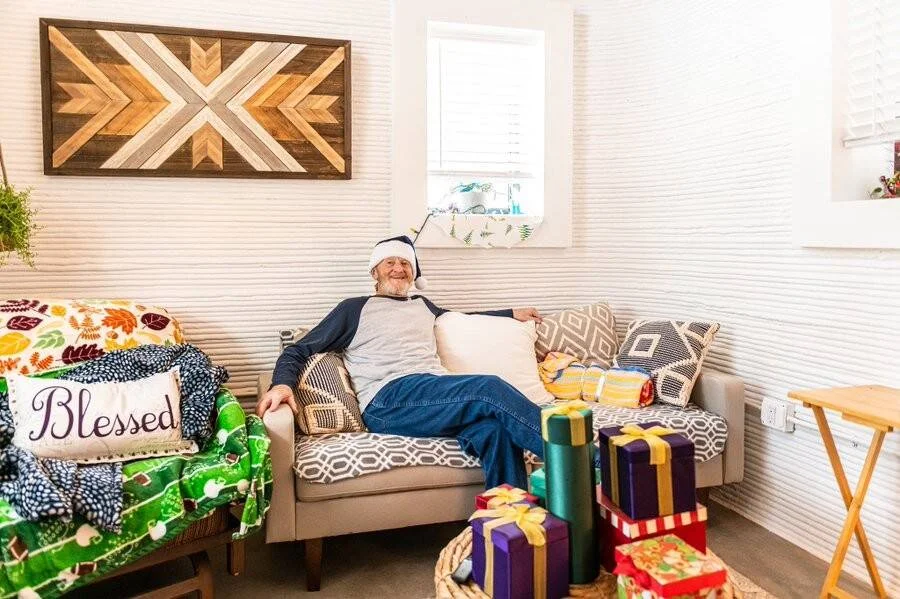 Hombre mayor sonriente con gorro de Navidad recostado en un sofá rodeado de regalos envueltos en sala decorada.