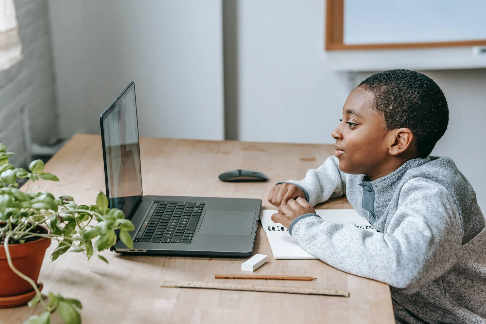 Niño concentrado mirando la pantalla de una laptop mientras tiene un cuaderno y útiles escolares sobre el escritorio.