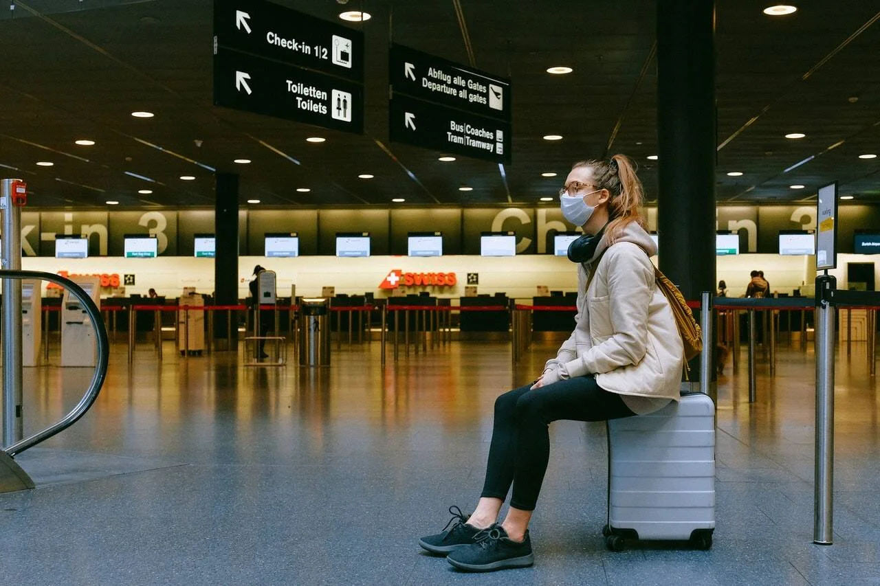 Mujer con mascarilla sentada sobre su maleta en un aeropuerto casi desierto, frente a los mostradores de check-in de Swiss.