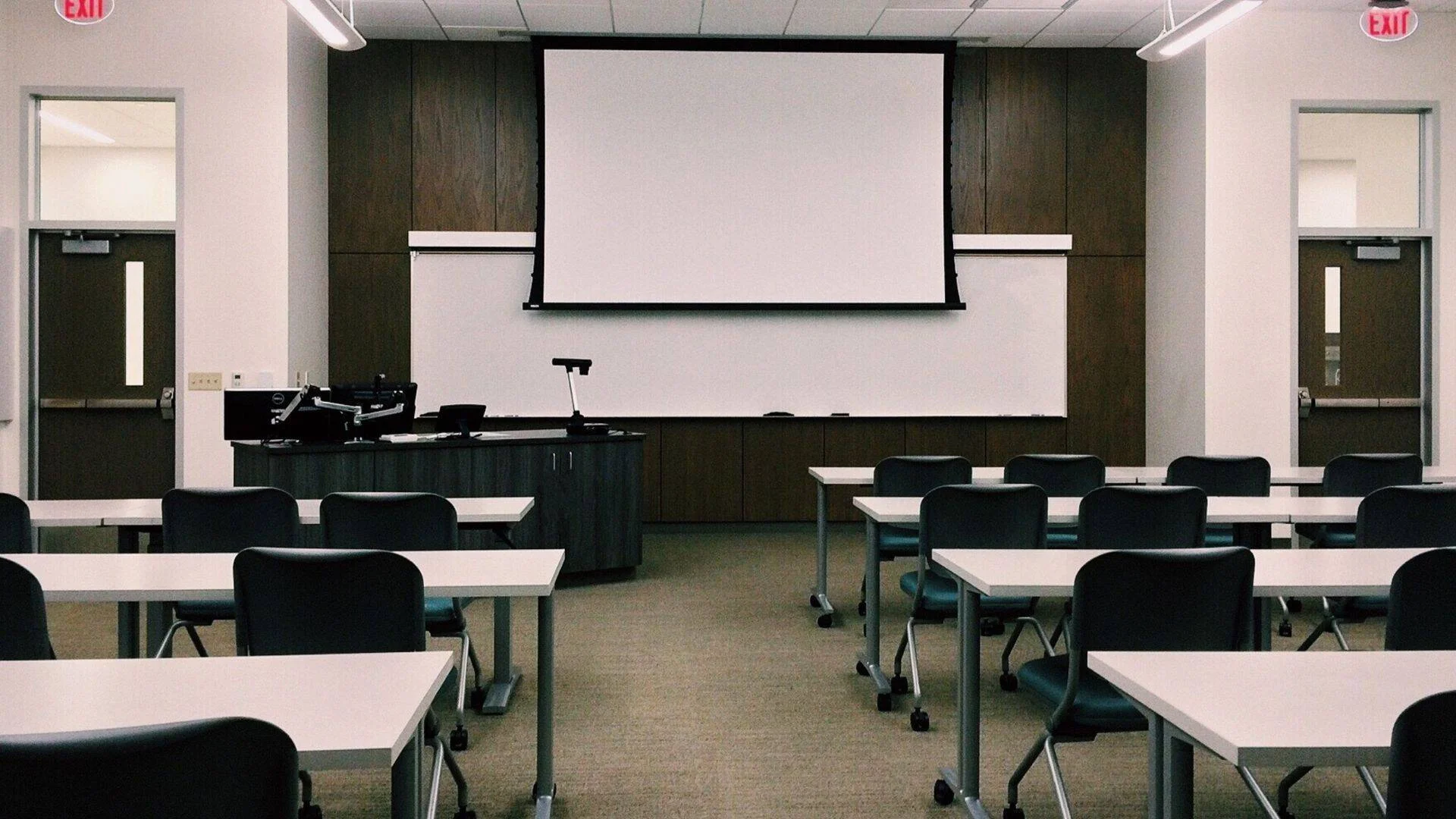 Aula vacía con filas de mesas blancas, sillas oscuras y una pantalla de proyección desplegada al frente del salón.
