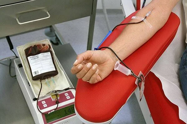Brazo de una persona con vía intravenosa durante una donación de sangre, junto a una bolsa de sangre sobre una balanza.