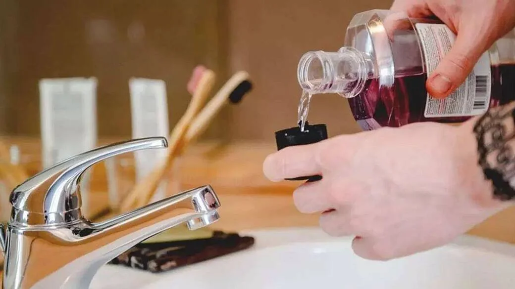 Manos sirviendo enjuague bucal de color morado en un vasito, junto a un lavabo con grifo y cepillos de dientes de bambú al fondo.