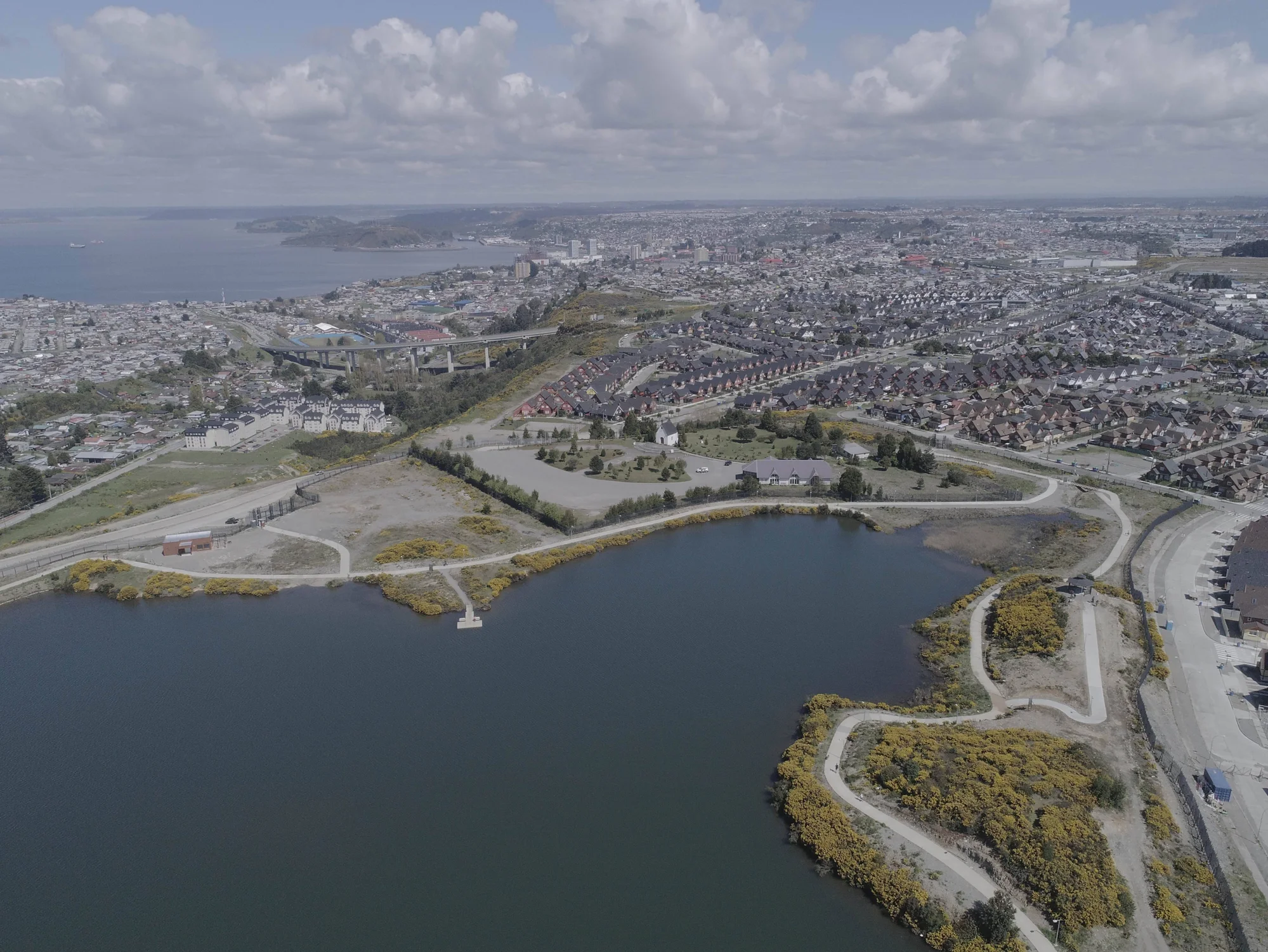 Panorámica aérea de una ciudad costera con una laguna en primer plano, zonas residenciales y un puente al fondo.