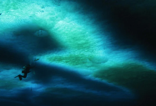 Un buzo nada bajo una superficie de hielo con tonos verdes y turquesas que filtran la luz desde arriba.