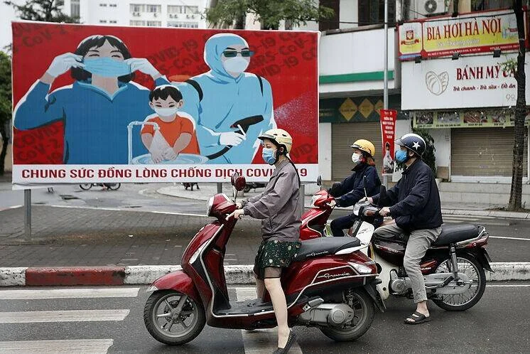 Personas con mascarilla en moto pasan frente a un cartel de propaganda contra el COVID-19 en una calle de Vietnam.