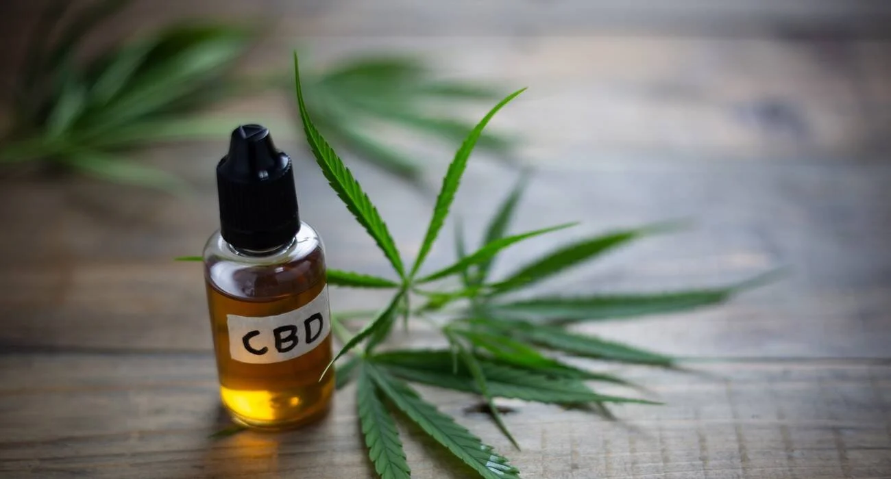 Frasco con gotero etiquetado como CBD, conteniendo aceite dorado, junto a hojas verdes de cannabis sobre superficie de madera.