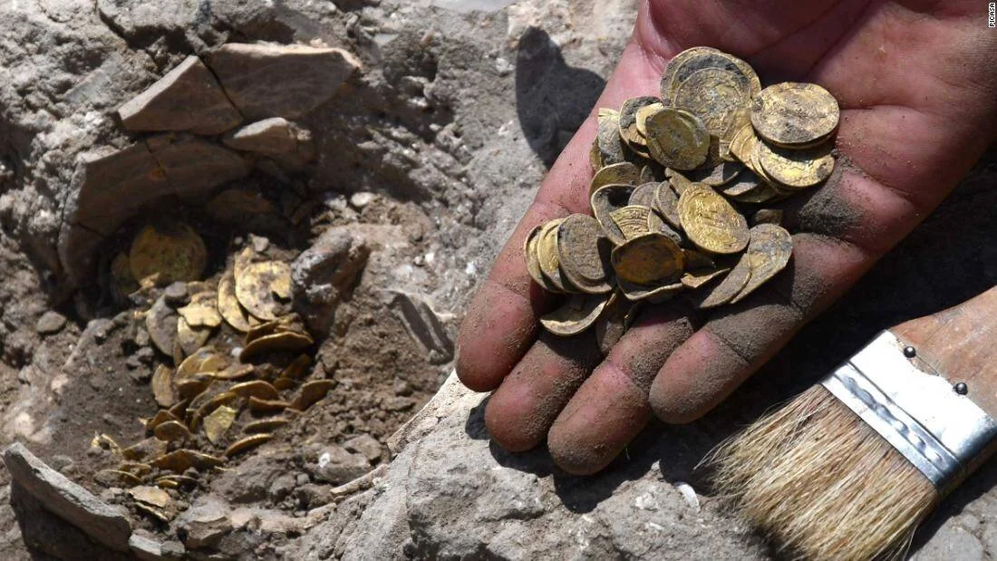 Mano sosteniendo decenas de monedas de oro antiguas junto a una vasija rota enterrada en el suelo durante una excavación arqueológica.