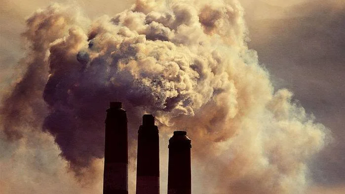 Tres chimeneas industriales en silueta expulsan densas nubes de humo gris al cielo, representando contaminación atmosférica.
