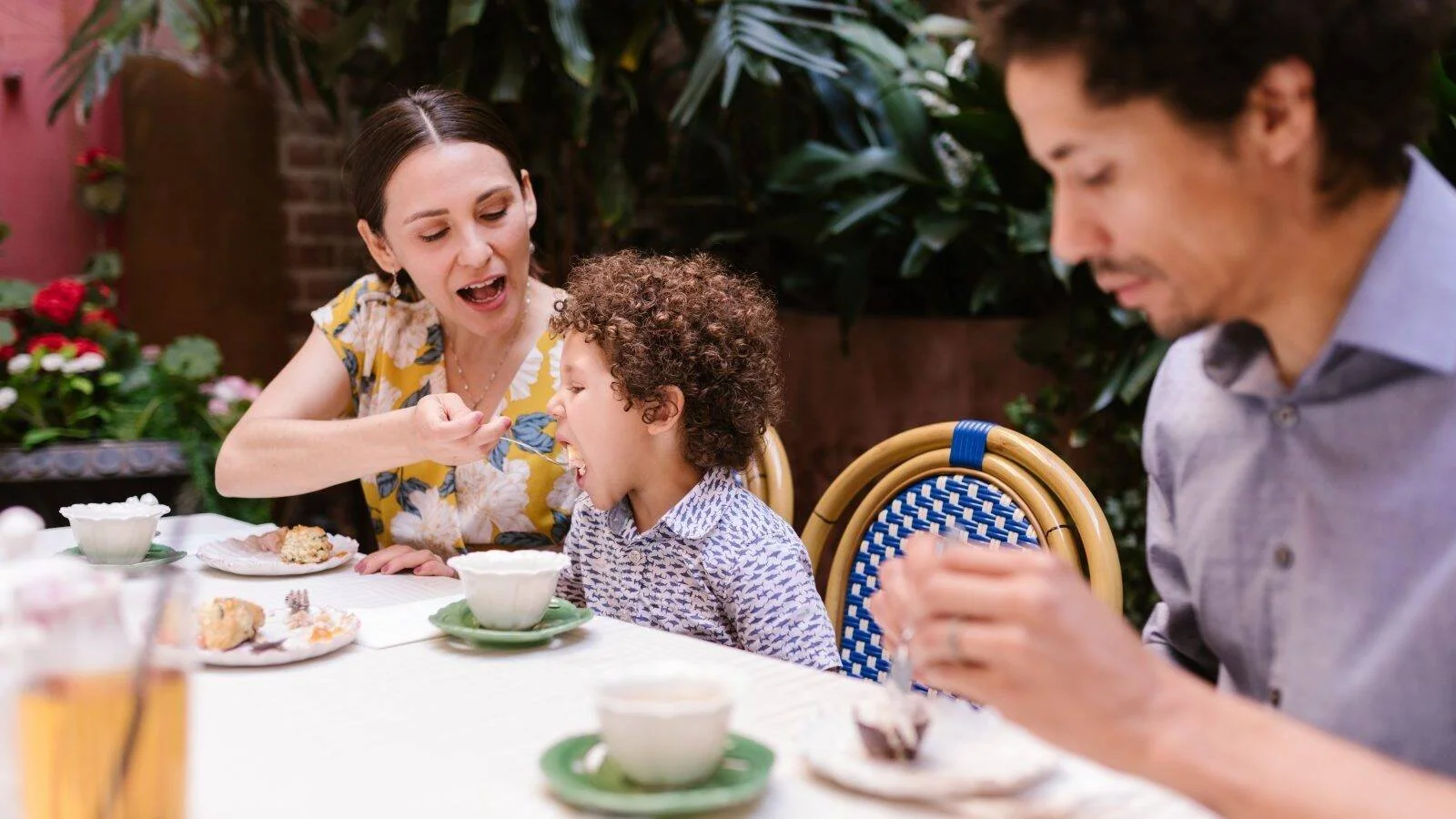 Una madre da de comer con una cuchara a su hijo pequeño rizado mientras el padre come junto a ellos en un restaurante con plantas.