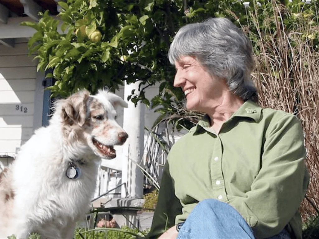 Mujer de cabello gris sonríe mirando a un perro blanco con manchas marrones sentados juntos en un jardín.