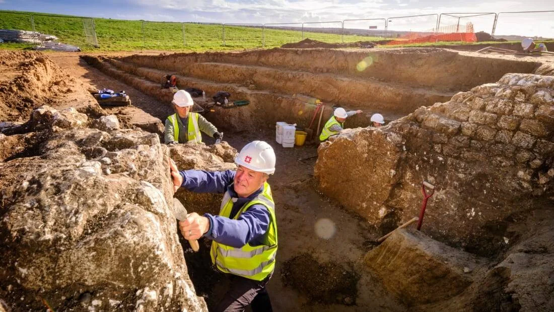 Varios arqueólogos con cascos y chalecos reflectantes excavan una trinchera con restos de muros de piedra en un campo abierto.