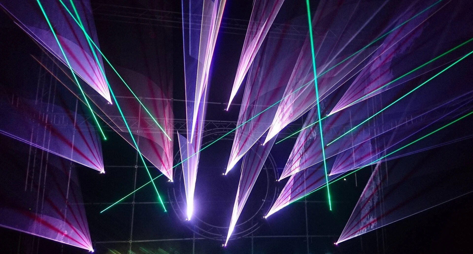 Haces de luz láser verde, rosa y violeta proyectados hacia arriba en un espectáculo nocturno con estructuras metálicas.