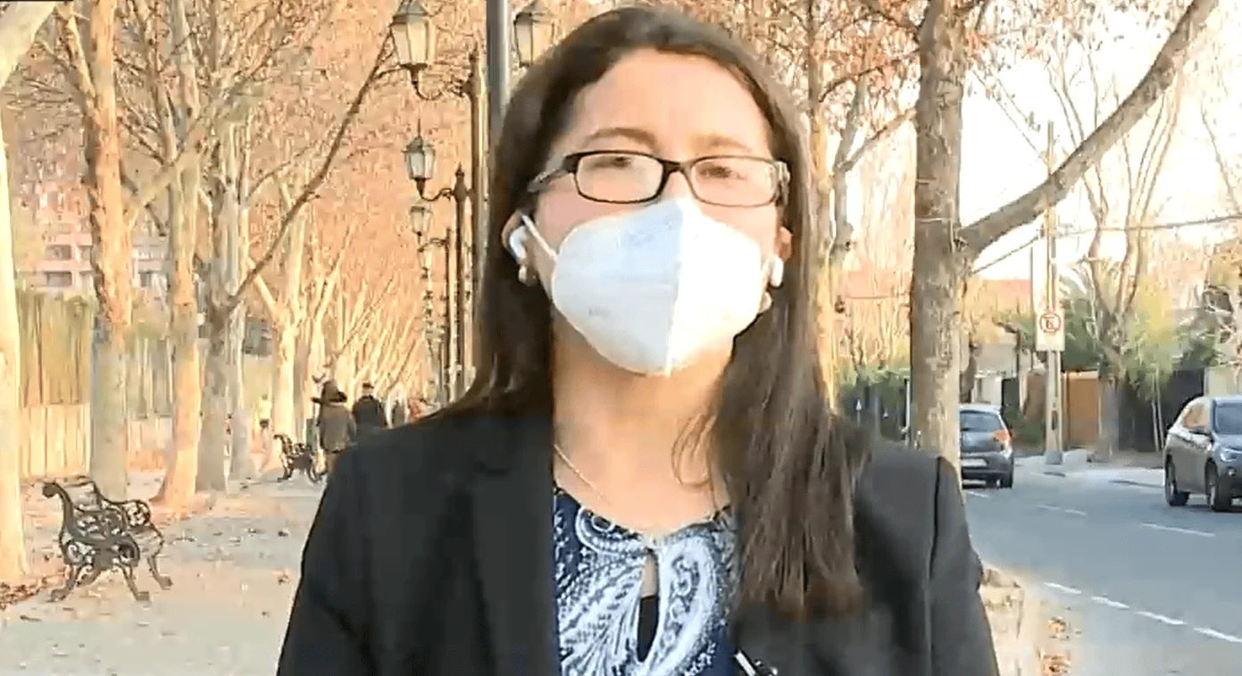 Mujer con gafas y mascarilla FFP2 blanca en un paseo arbolado, con auriculares inalámbricos y chaqueta oscura.