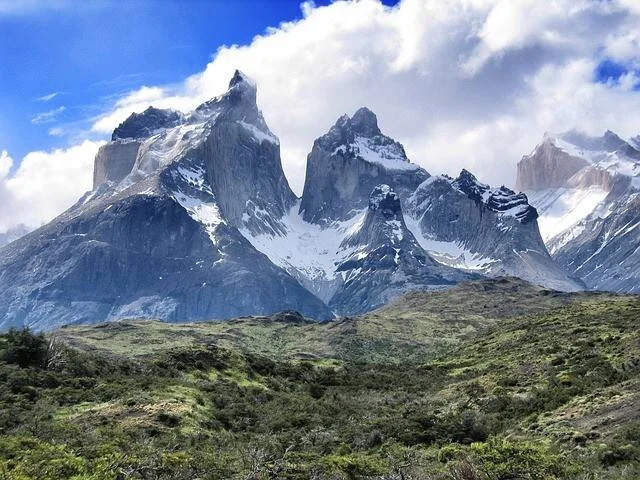 Imponentes picos rocosos nevados de Torres del Paine se elevan sobre una pradera verde bajo cielo parcialmente nublado.