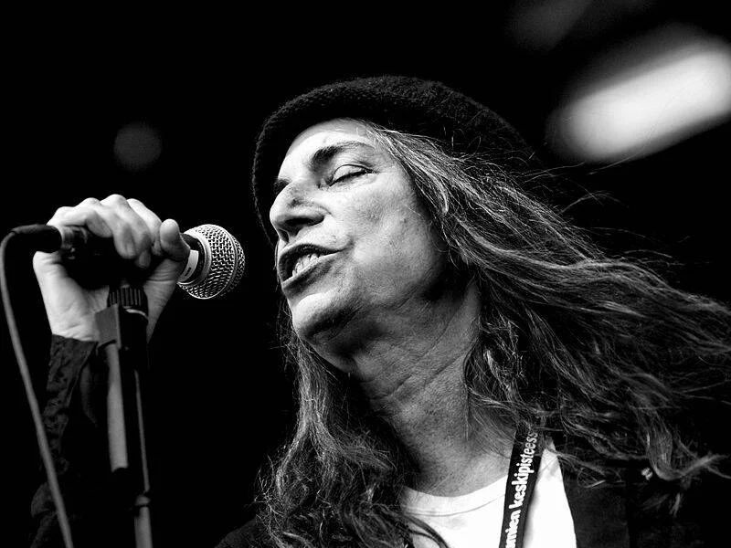 Cantante de cabello largo y gorro negro interpreta una canción frente al micrófono en un concierto, fotografía en blanco y negro.