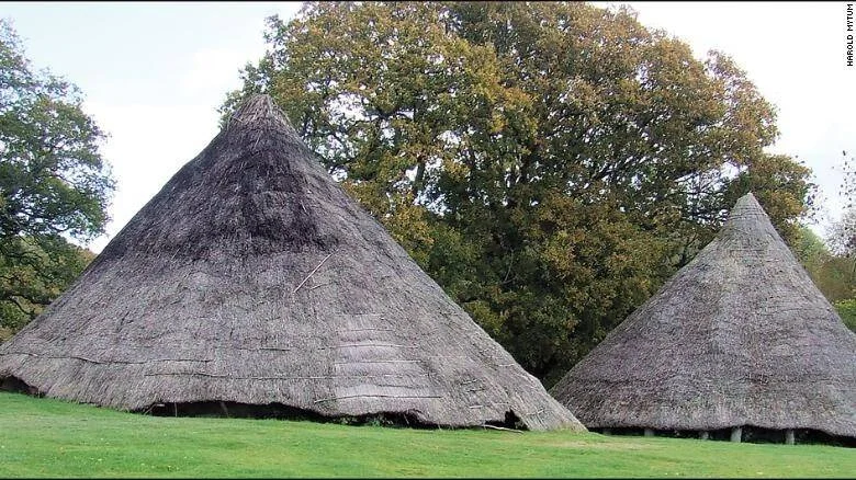 Dos chozas de época prehistórica con techos cónicos de paja, rodeadas de césped verde y árboles frondosos.