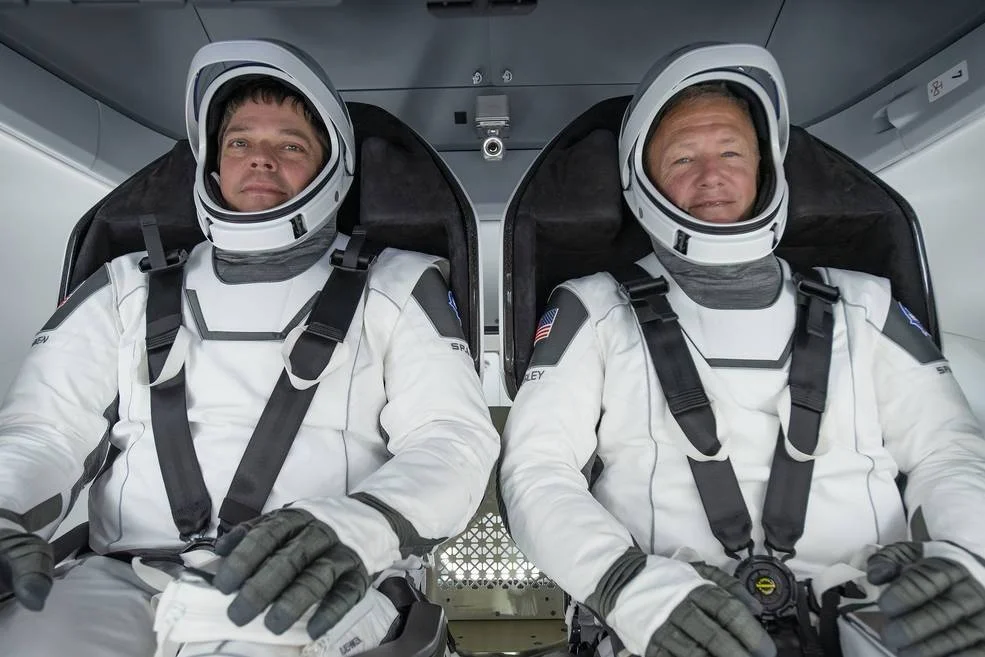 Dos astronautas con trajes blancos de SpaceX y cascos abiertos sentados en la cápsula Dragon, listos para el lanzamiento.