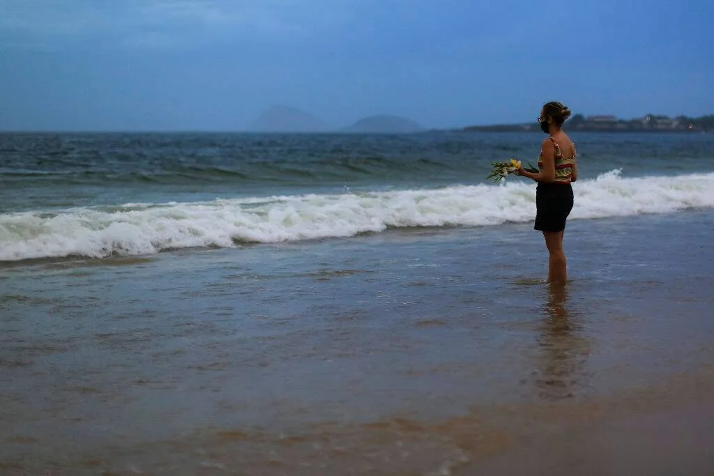 Mujer de pie en la orilla del mar sosteniendo un ramo de flores amarillas, lista para ofrendarlo al océano.