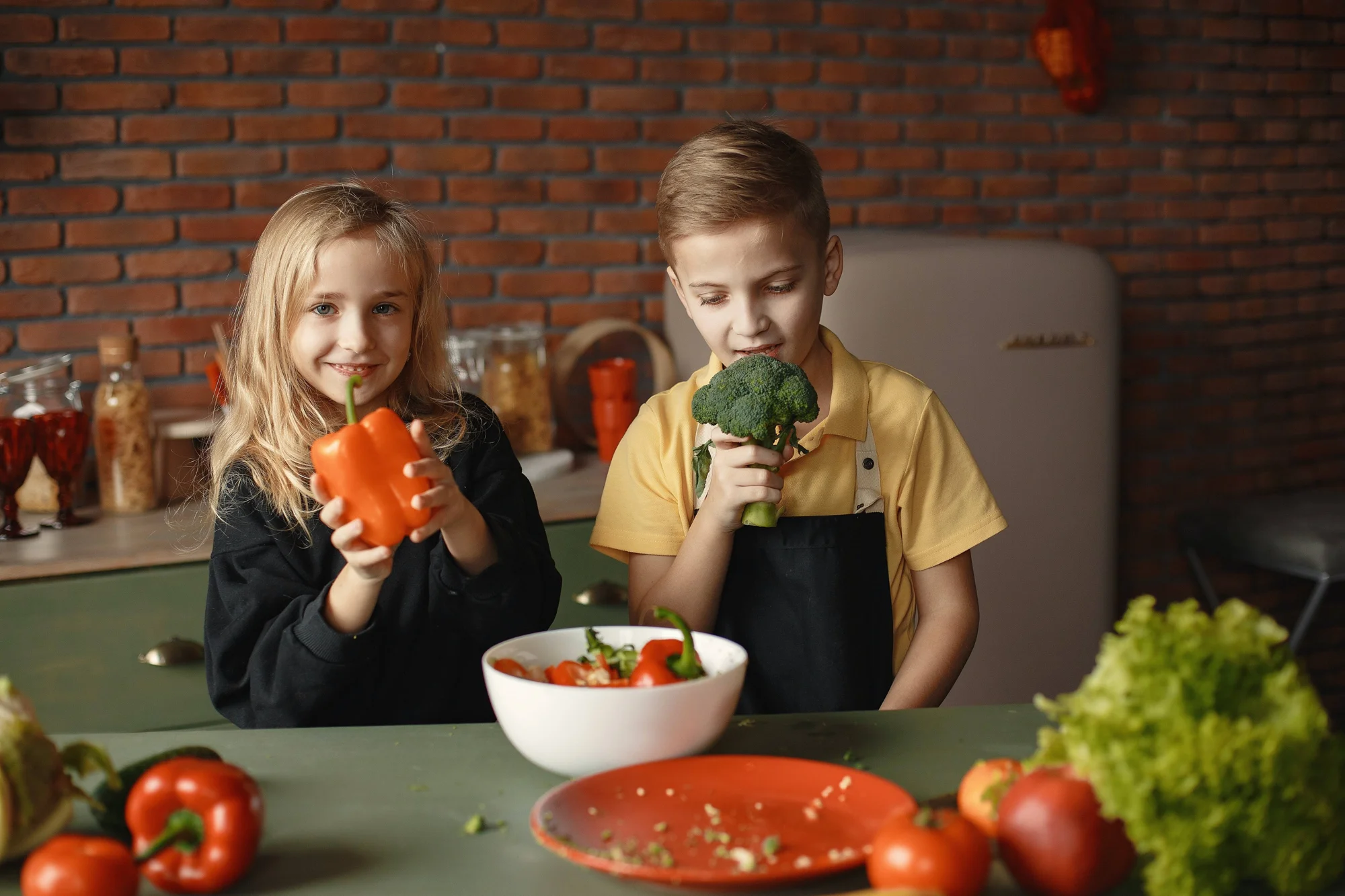 Dos niños sostienen un pimiento rojo y brócoli junto a una mesa llena de verduras frescas en una cocina.