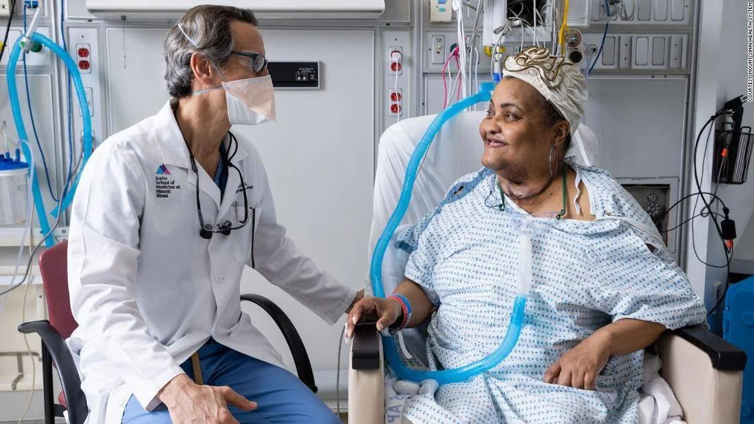 Un médico del Mount Sinai habla con una paciente sentada en silla hospitalaria conectada a tubos de soporte vital.