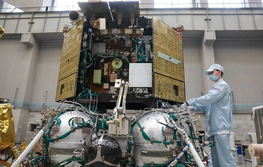 Un técnico con traje de sala limpia y mascarilla revisa un satélite de grandes paneles dorados en una instalación aeroespacial.