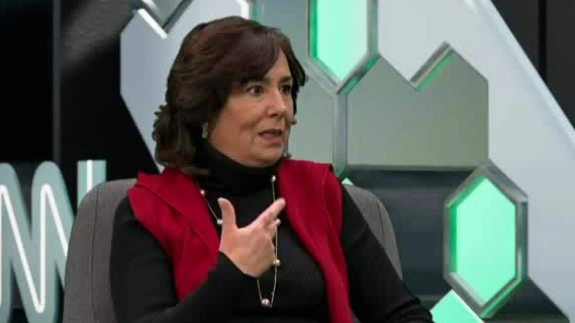 Mujer de cabello oscuro con chaleco rojo gesticula mientras habla en un estudio de televisión con decorado hexagonal verde.