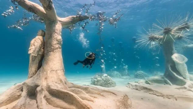 Un buzo nada entre esculturas de árboles y figuras humanas en el fondo marino de aguas cristalinas.