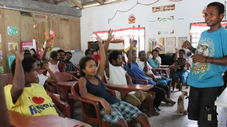 Grupo de personas levantando la mano durante una reunión o taller comunitario en una sala modesta.