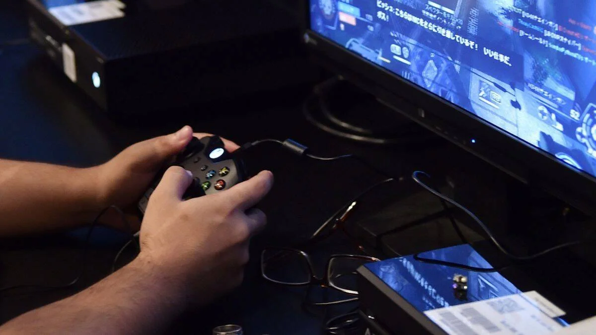 Manos sosteniendo un control Xbox negro mientras juega un videojuego con texto en japonés en la pantalla.