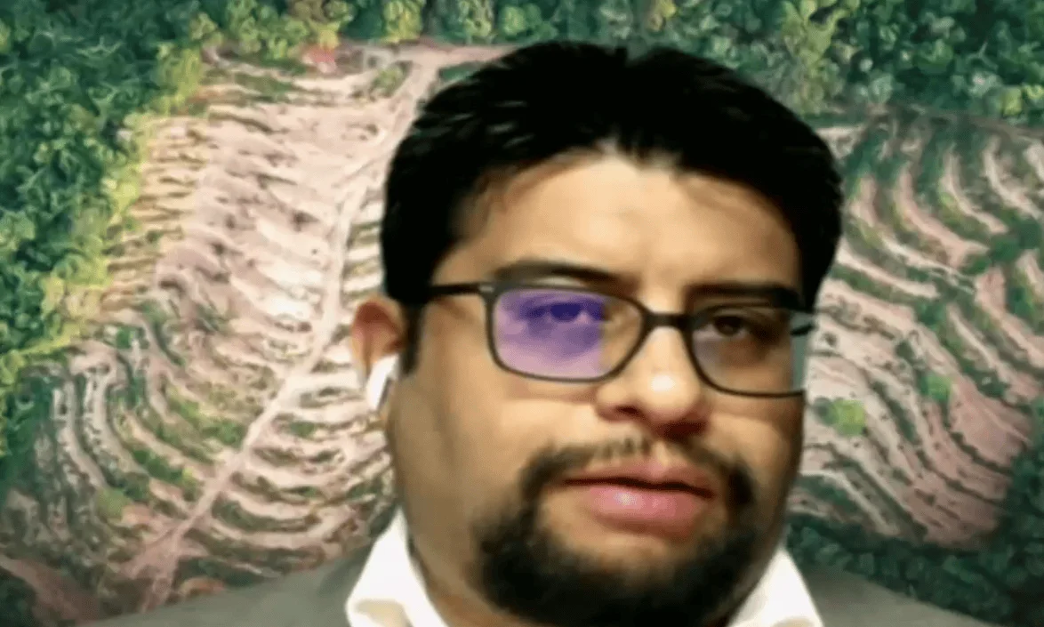 Hombre joven con lentes y auricular inalámbrico frente a un fondo con imagen aérea de zona deforestada.