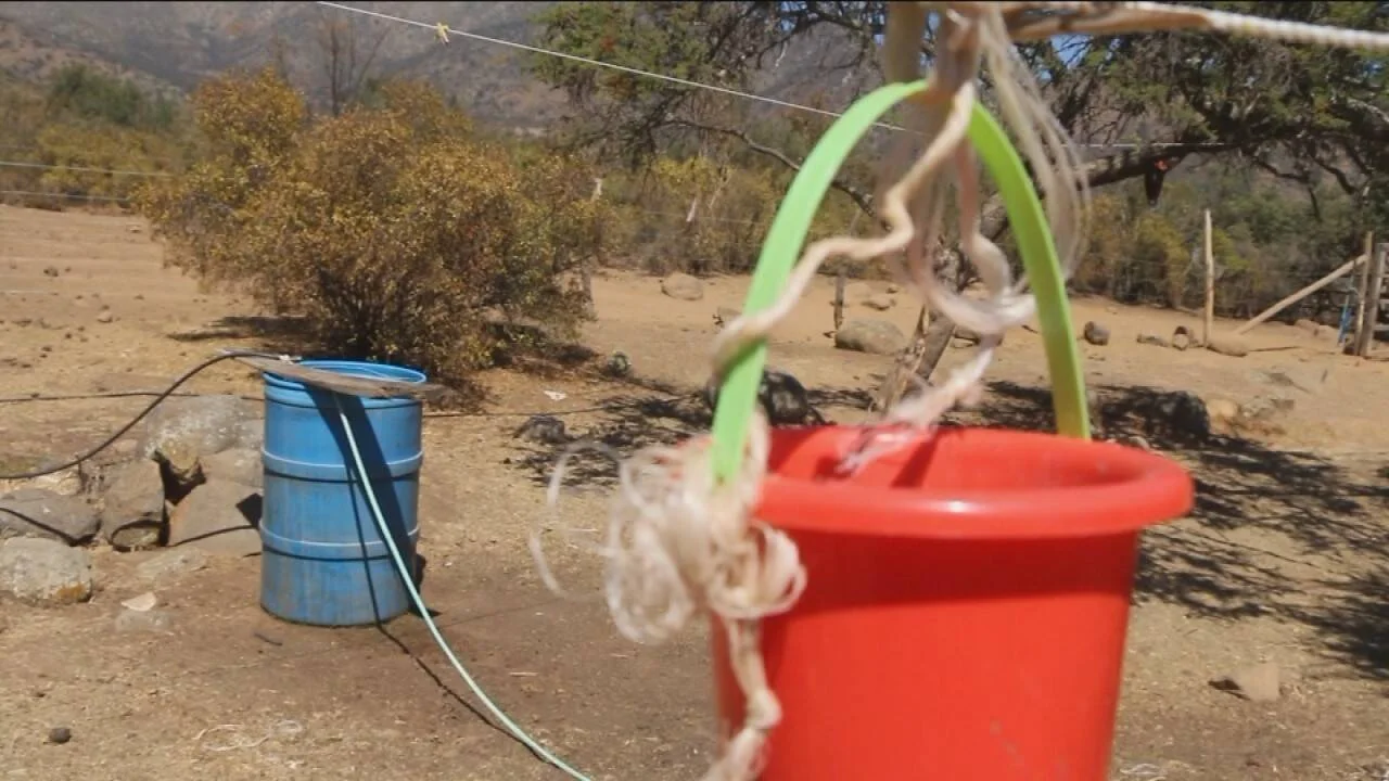 Balde rojo colgado con cuerda y tambo azul con manguera en terreno seco y árido con montañas al fondo.