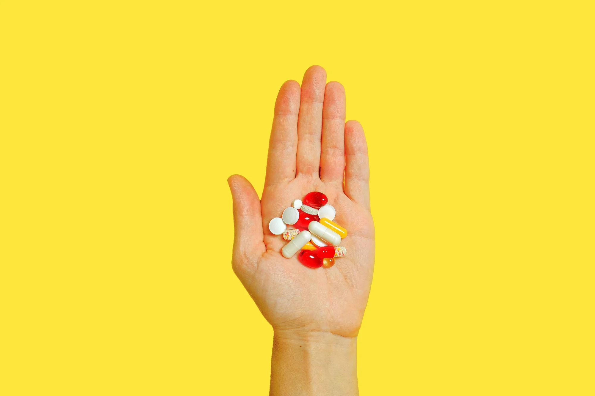 Una mano abierta sostiene un conjunto de pastillas y cápsulas de distintos colores sobre fondo amarillo.