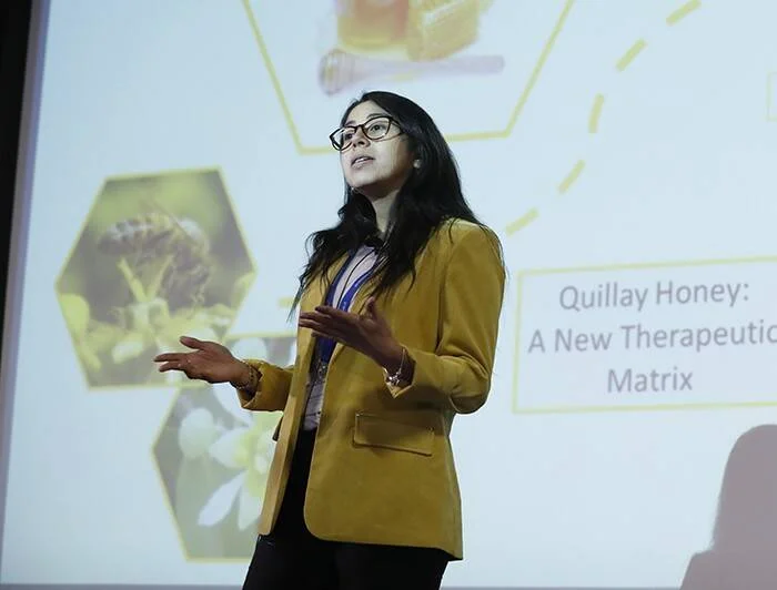 Mujer con blazer amarillo presenta una ponencia sobre 'Quillay Honey: A New Therapeutic Matrix' ante una pantalla de proyección.