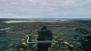Vista aérea de un puente peatonal sobre una grieta en campo de lava volcánica oscura, con el océano al fondo bajo cielo nublado.