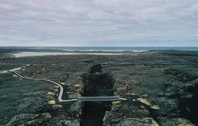 Vista aérea de un puente peatonal sobre una grieta en campo de lava volcánica oscura, con el océano al fondo bajo cielo nublado.