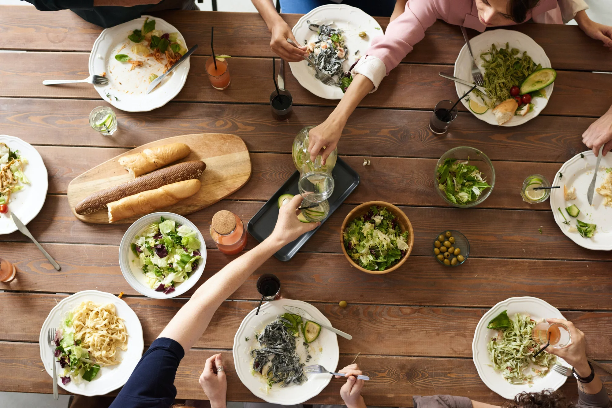 Vista cenital de varias personas compartiendo platos de pasta, ensaladas y pan en una mesa de madera.