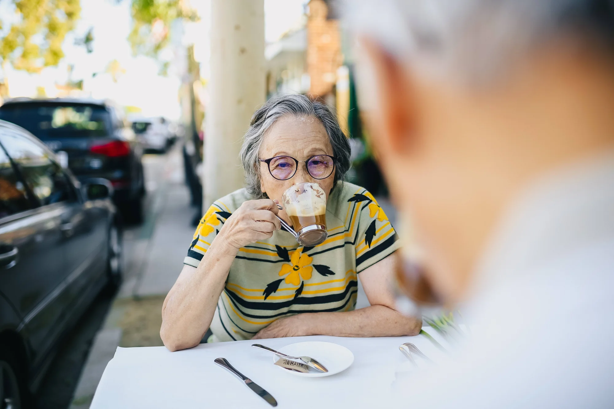 Mujer mayor con gafas redondas bebe un café con crema en la terraza de un bar, sentada frente a otra persona.