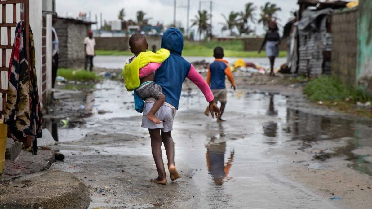 Un niño carga a otro en brazos mientras camina descalzo por una calle anegada en una zona humilde afectada por inundaciones.