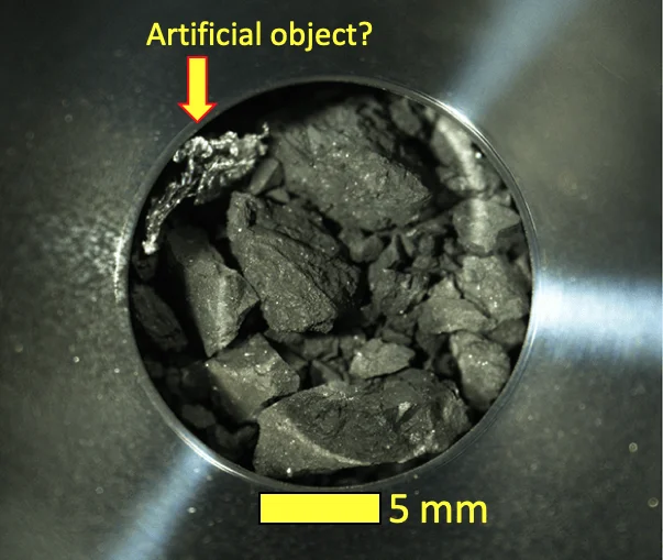 Fragmentos de roca oscura en recipiente metálico con flecha señalando un posible objeto artificial; escala de 5 mm.