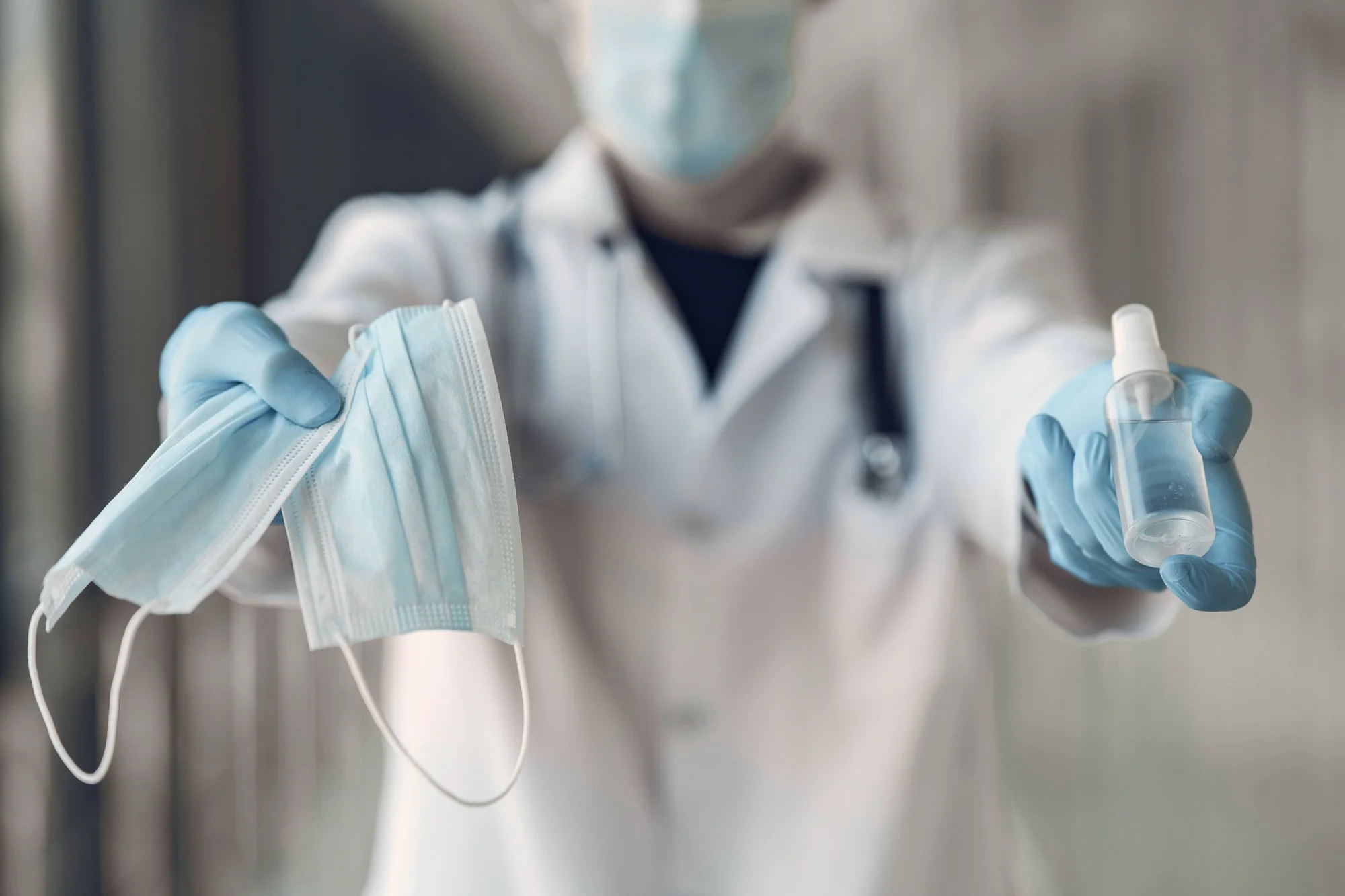 Personal sanitario con guantes azules extiende mascarillas quirúrgicas en una mano y un frasco de gel desinfectante en la otra.