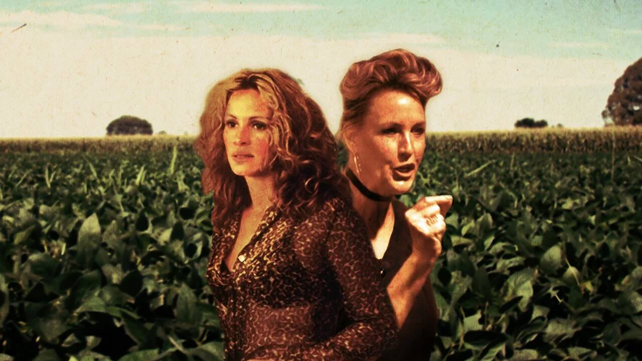 Dos mujeres de cabello rojizo conversan en medio de un extenso campo de cultivo verde bajo un cielo despejado.