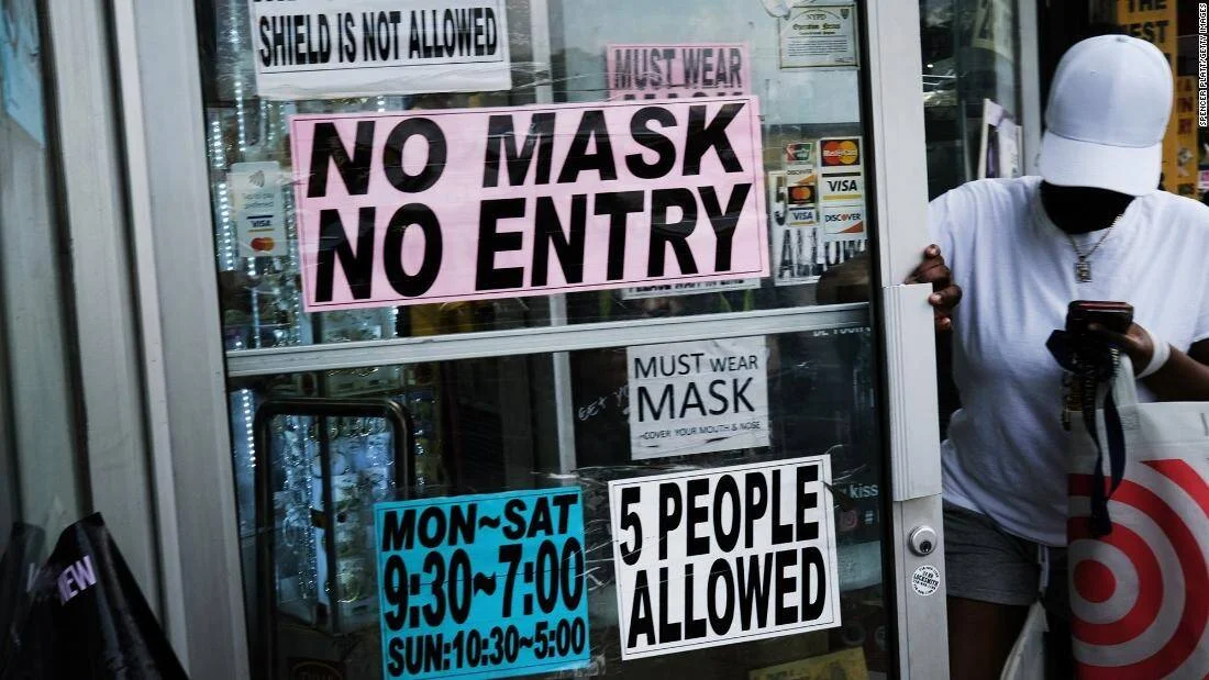Persona entrando a una tienda con carteles que exigen mascarilla y limitan el aforo a 5 personas durante la pandemia.