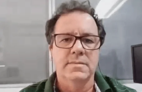 Hombre de mediana edad con anteojos de marco rojo y campera verde grabado en video en una oficina.