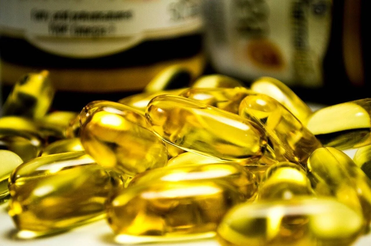 Montón de cápsulas de gel doradas de suplemento vitamínico con frascos desenfocados al fondo.