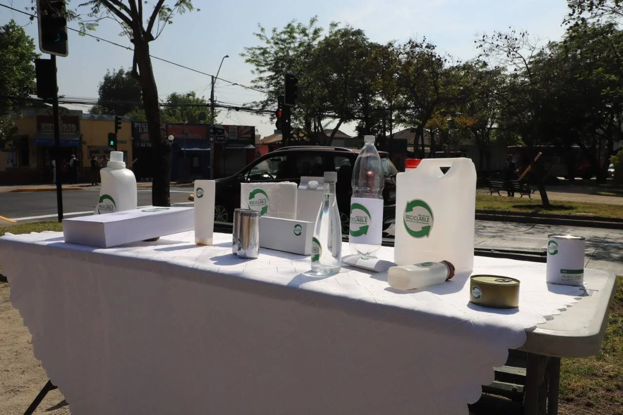 Mesa con diversos envases etiquetados como reciclables —botellas, latas, cajas y bidones— expuestos al aire libre en una plaza urbana.