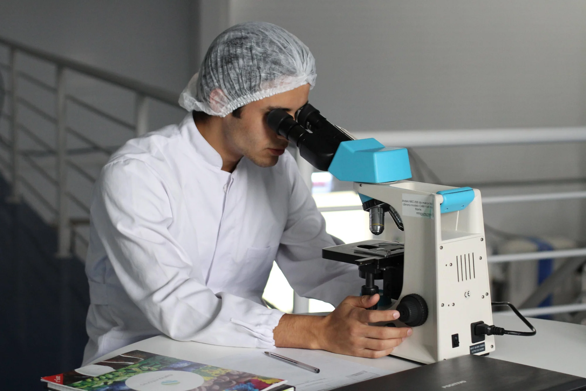 Técnico de laboratorio con cofia y bata blanca observa una muestra a través de un microscopio óptico.