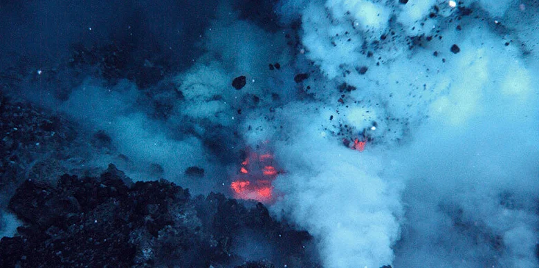 Vista aérea nocturna de una erupción volcánica con lava incandescente, rocas proyectadas y densa nube de humo.